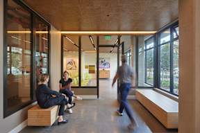Three people are sitting on a bench in a hallway with windows on one side.