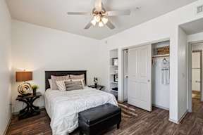 One Bedroom Apartments in West Phoenix, AZ - Del Mar - a Bedroom with a White Bed, a Bedside Table with a Lamp, Shelving Built Into the Wall, a Closet with Sliding Wood Doors, Wood-Style Flooring, and a Ceiling Fan