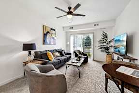 A living room with a grey couch, a grey armchair, a wooden coffee table, a ceiling fan, a flat screen TV, and a painting on the wall.