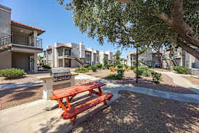 A red picnic table is in the middle of a gravel area with houses in the background.