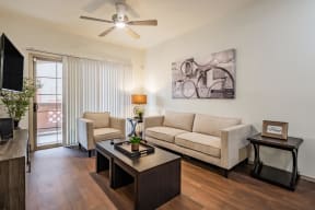 Apartments in West Phoenix AZ - Del Mar - Living Room with Couch, Coffee Table, Chair, End Table, Lamp, Ceiling Fan, TV Stand, and Large Sliding Glass Door Leading to Balcony/Patio.