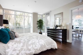 One-Bedroom Apartments in Irvine, CA - Astoria at Central Park West - Bedroom with Dark Wood-Tone Vinyl Flooring, Bed, Wooden Dresser, Framed Mirror, Lamps, House Plant, and Large Floor-to-Ceiling Windows.