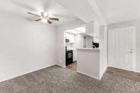 A room with a ceiling fan and a kitchen area in the background.