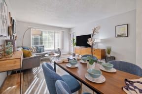 Hayward, CA, Apartments - Glen Oaks - Dining Area with Table and Plush Chairs Connected to Living Room with Gray Couch, Small Round Coffee Table, TV Table with Cabinets, Wood-Inspired Flooring, and a Large Window with White Blinds.