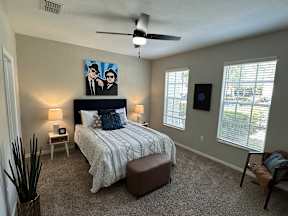 A bedroom with a bed, a ceiling fan, and a painting of two people.