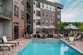 A swimming pool surrounded by chairs and umbrellas in front of a multi-story apartment building.