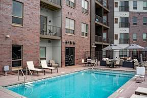 A pool surrounded by chairs and umbrellas in front of a brick building.