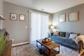 A living room with a grey couch, a wooden coffee table, and a large window with white blinds.