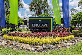 A sign for Enclave Apartments Homes is surrounded by flowers and flags.