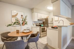 Apartments Near Westgate, Glendale, AZ - Ardella on 67th - Kitchen with Open Space to Dining Area, Featuring Stainless Steel Appliances, White Cabinetry, a Table, and Chairs.