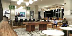A modern office space with a long wooden table in the center and people sitting and standing around it.