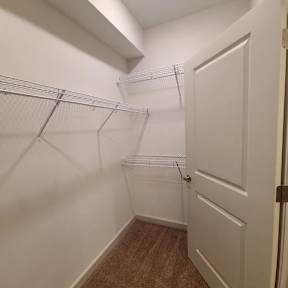 A white door is on the right side of a hallway with a carpeted floor and a wall-mounted shelf.