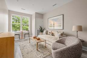 A living room with a grey sofa, a wooden table, a grey armchair, a wooden cabinet, a rug, a potted plant, a painting, a window, and a lamp.