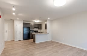 an empty living room with a kitchen with a black refrigerator