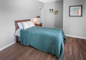 A bed with a blue blanket is in the center of a room with wood flooring and white walls.