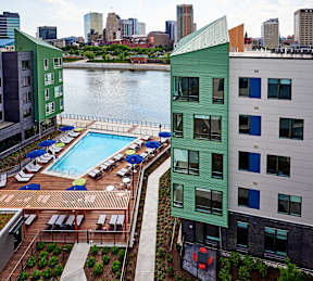 A green building with a pool in the foreground and a city skyline in the background.