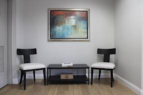 A painting hangs on a wall above a black coffee table with two chairs.