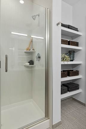 A white bathroom with a glass shower door and shelves with towels and other bathroom accessories.