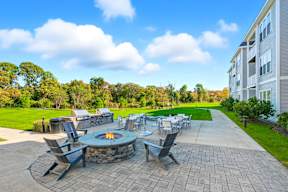 A patio with a fire pit and chairs is surrounded by a grassy area and a building.