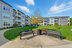 A park area with benches and a tree in front of apartment buildings.