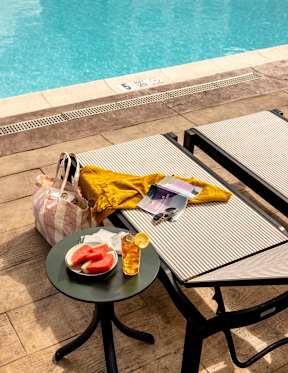 A table with a plate of watermelon and a drink sits next to a pool.