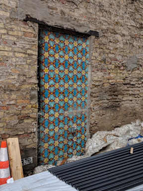 A door with a pattern of blue and orange diamonds is set in a brick wall.