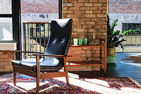 A black leather chair with wooden legs sits on a rug in front of a brick wall.