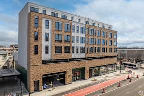 A modern building with a mix of brick and panel construction is situated on a busy street corner.