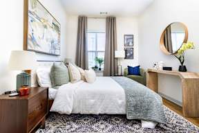A bedroom with a large bed, a nightstand with a lamp, a side table with a vase of flowers, and a mirror on the wall.