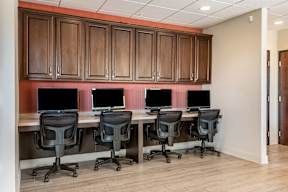 A row of four computers are on a desk with black chairs.