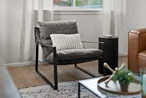 A grey chair with a white pillow is in the foreground of a room with a black table and a brown leather chair.
