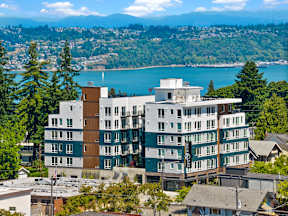 A large white building with a brown roof is in the foreground of a scenic view of a lake and mountains.
