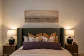 A bed with a green headboard and purple pillows is flanked by two lamps on wooden nightstands.