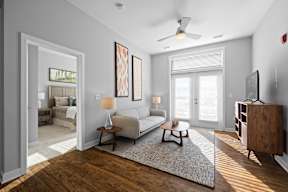 A spacious living room with hardwood-style flooring, double doors to a patio, and a ceiling fan near an doorway to a bedroom.