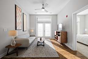 A living room with double doors to a patio providing natural light, hardwood-style flooring, a ceiling fan, and access to a bedroom.