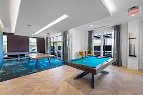 A clubhouse with a billiards table, a ping pong table, and a shuffleboard near doors leading to an outdoor community lounge.