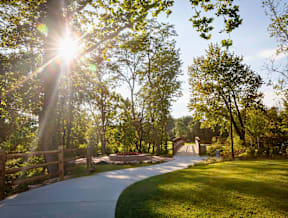 A beautiful trail with the sun shining through large trees and well-maintained grass leading to a bridge.
