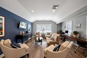 A clubhouse with two windows, hardwood-style flooring, comfortable seating areas, and a flat-screen TV.