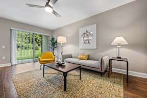 A living room with hardwood-style flooring, ceiling fan, and large sliding glass doors to private patio with well-maintained green space.