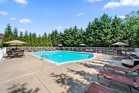 A gated resort-style pool with lounge chairs, tables with umbrellas, and a sundeck at Severgn Apartments.