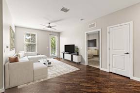 Bright living room at The Flats at Tioga Town Center with L-shaped sofa, TV console, ceiling fan, and balcony door.