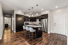 Modern kitchen with island, dark cabinetry, stainless appliances, three pendant lights, and dark wood floors at The Flats at Tioga Town Center.