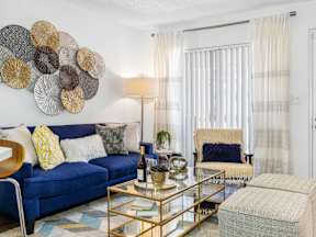 A living room with a blue couch, a glass coffee table, and a large decorative piece on the wall.