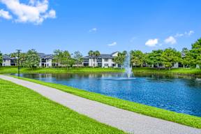 A serene landscape with a fountain in the middle of a lake surrounded by green grass and buildings in the background.