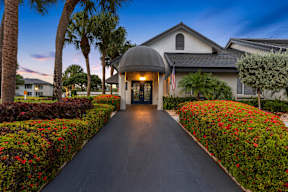 A house with a flag on the front porch is surrounded by red flowers and palm trees.