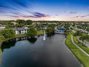 A serene lake with a fountain in the middle surrounded by houses and trees under a twilight sky.