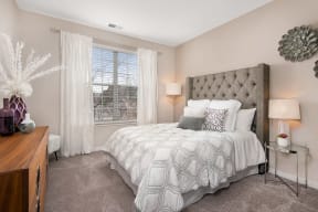 One-Bedroom Apartments in Naperville, IL - 803 Corday at Naperville - Bright Bedroom With Tufted Headboard, Plush Bedding, Soft Carpet, Large Window With Sheer Curtains, And Bedside Lamps.