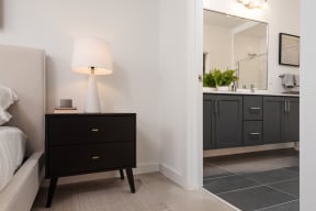 a bedroom with a black nightstand with a lamp and a bathroom with a sink