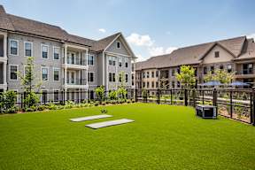A grassy area in front of apartment buildings with a trash bin and a bench.