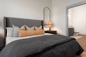 A bed with a grey headboard and a grey blanket with a lamp on the side table.